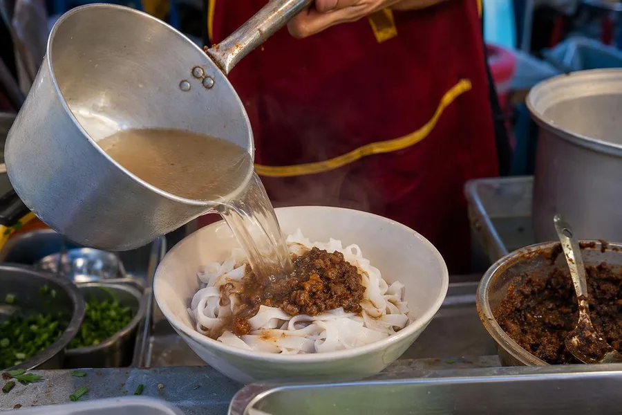 Heldere bouillon gaat over khao soi-noedels en vleessaus op de avondmarkt van Luang Namtha.