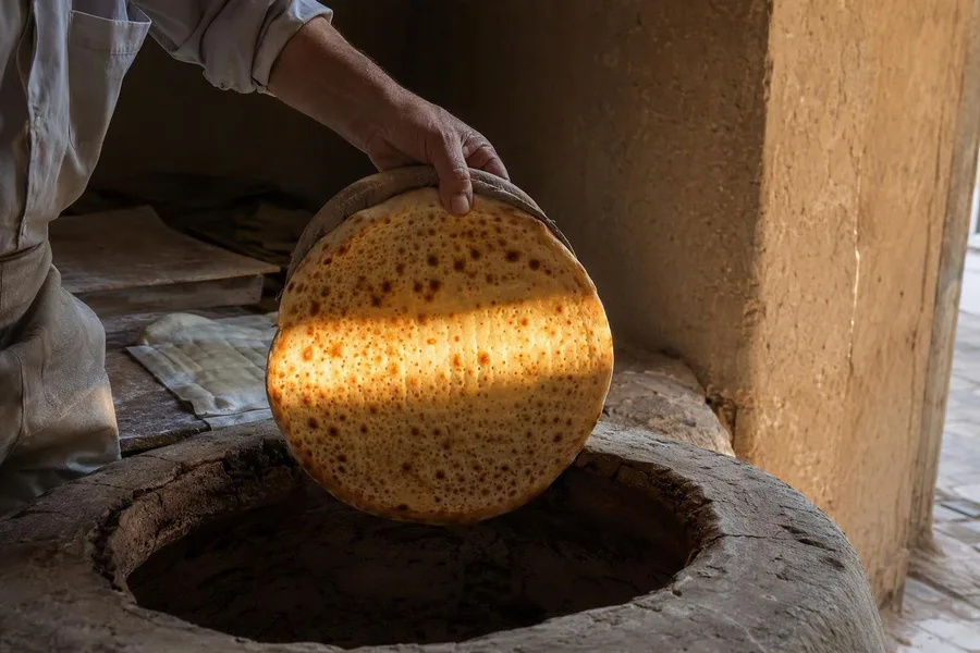 Een hand tilt verse taftoon uit de tanur in gefilterd licht bij de deuropening van de bakkerij in Yazd.