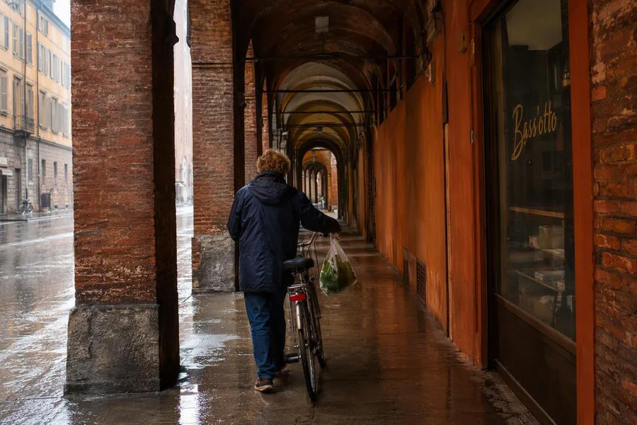 A cyclist with a grocery bag disappears beneath the porticoes near Bass’otto on Via Ugo Bassi.