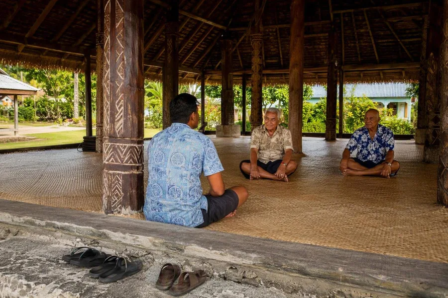 Een bezoeker zit met gekruiste benen in een dorpsfale op Upolu, met schoenen buiten.
