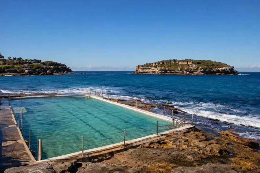 Zout water klotst tegen het Ross Jones Memorial Pool op de zuidelijke rotsen van Coogee, met Wedding Cake Island erachter.