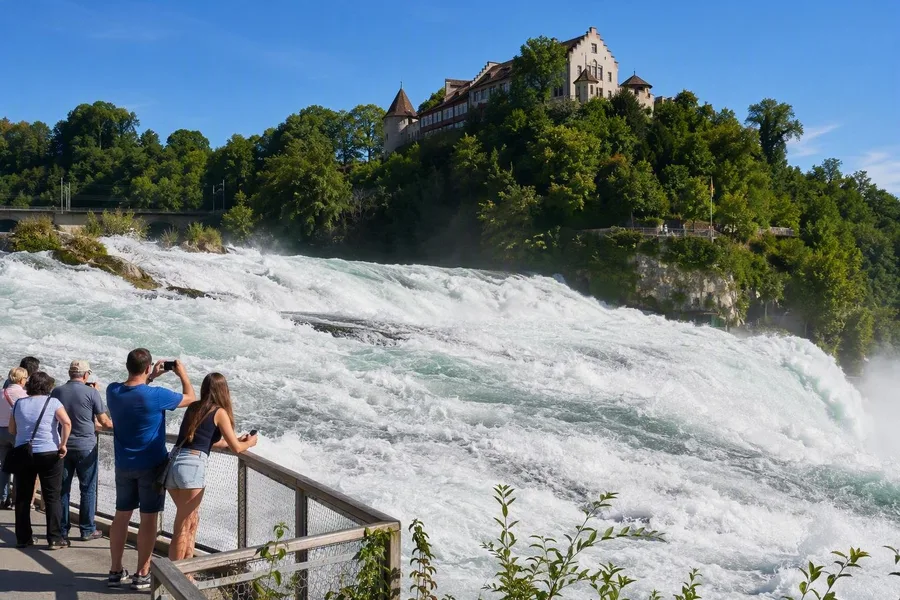 Bezoekers bekijken de Rijnwaterval vanaf een lager platform op de zuidoever, onder Schloss Laufen.