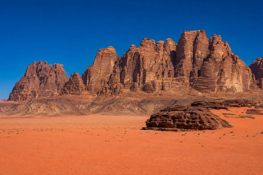 Gelaagde zandsteenmassieven rijzen op boven een vallei met roze zand in Wadi Rum.