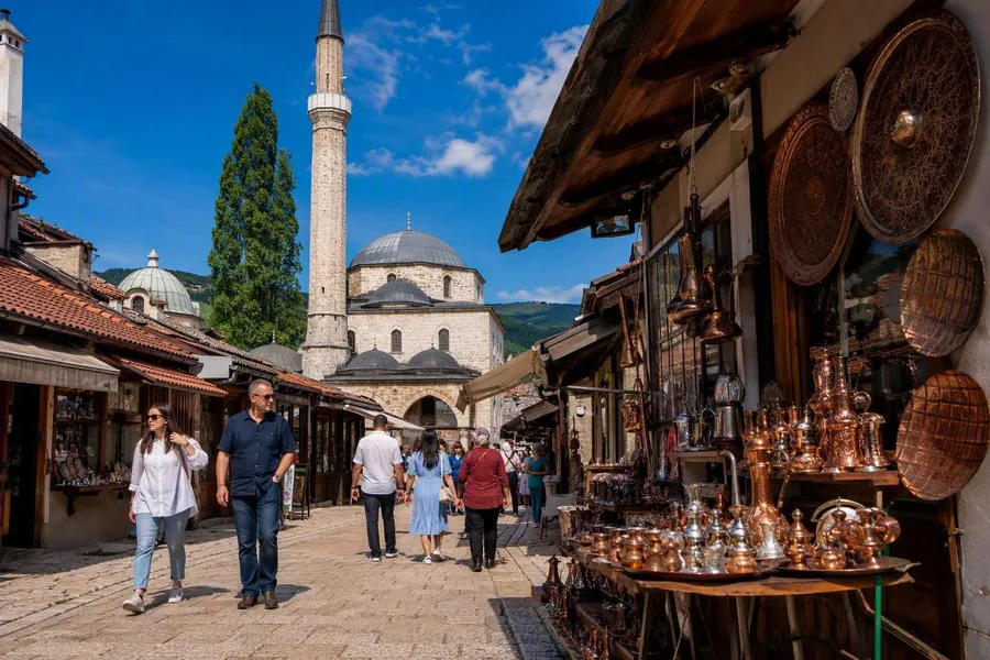 Voetgangers lopen langs koperwinkels in Sarajevo’s werkende Baščaršija-bazaar, bij de Gazi Husrev-begmoskee.