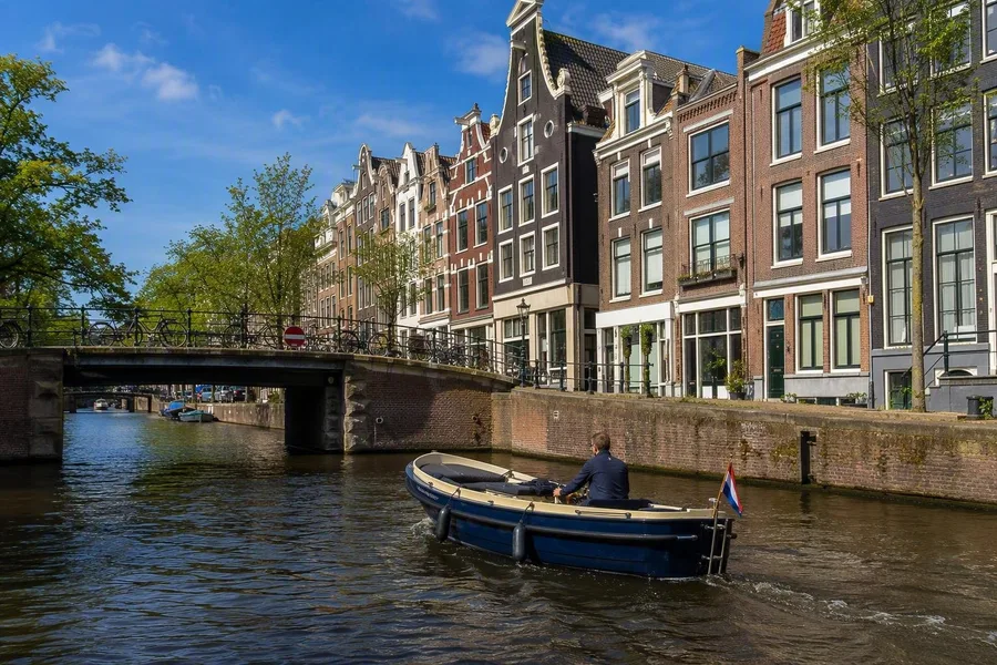 Een kleine elektrische boot vaart onder een brug langs Amsterdamse grachtenpanden met gevels.